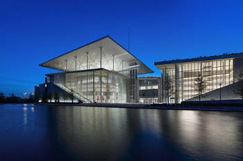 Photo taken at night showing a large illuminated glass building with a lake in front of it